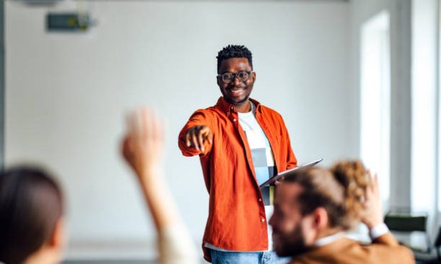 Handsome cheerful man in a orange shirt standing in front of an audience holding a tablet and using hand gestures to interact with the audience.